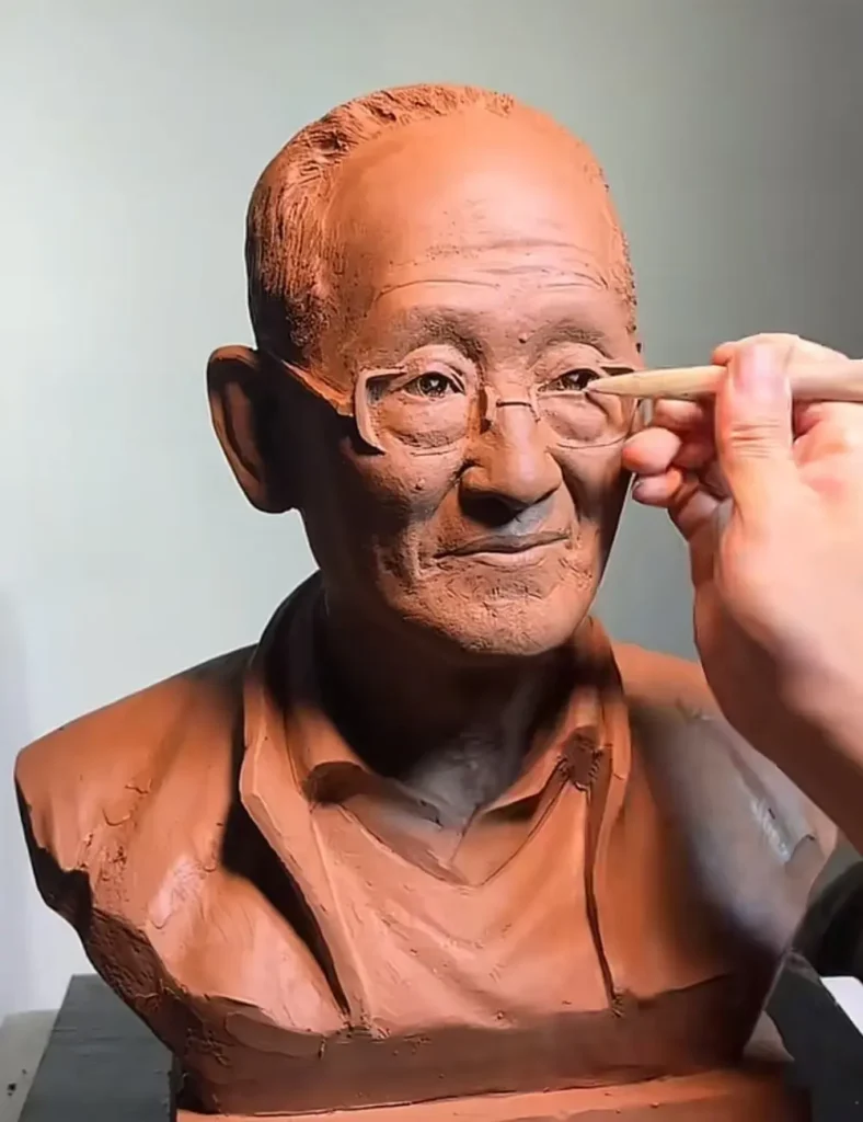 The custom memorial bronze bust for an elderly man with glasses, mid-creation, showcasing the sculptor’s work on his clothing and facial details.