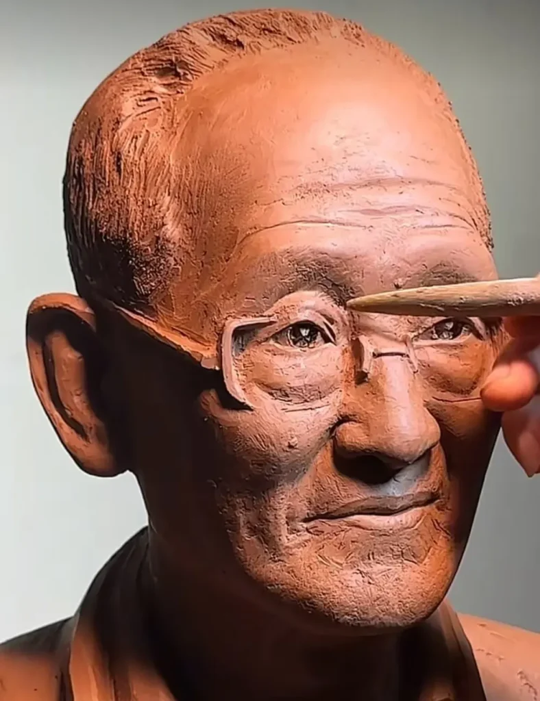 The sculptor at work on the custom memorial bronze bust for an elderly man with glasses, carefully shaping the glasses and facial features to match his photo.