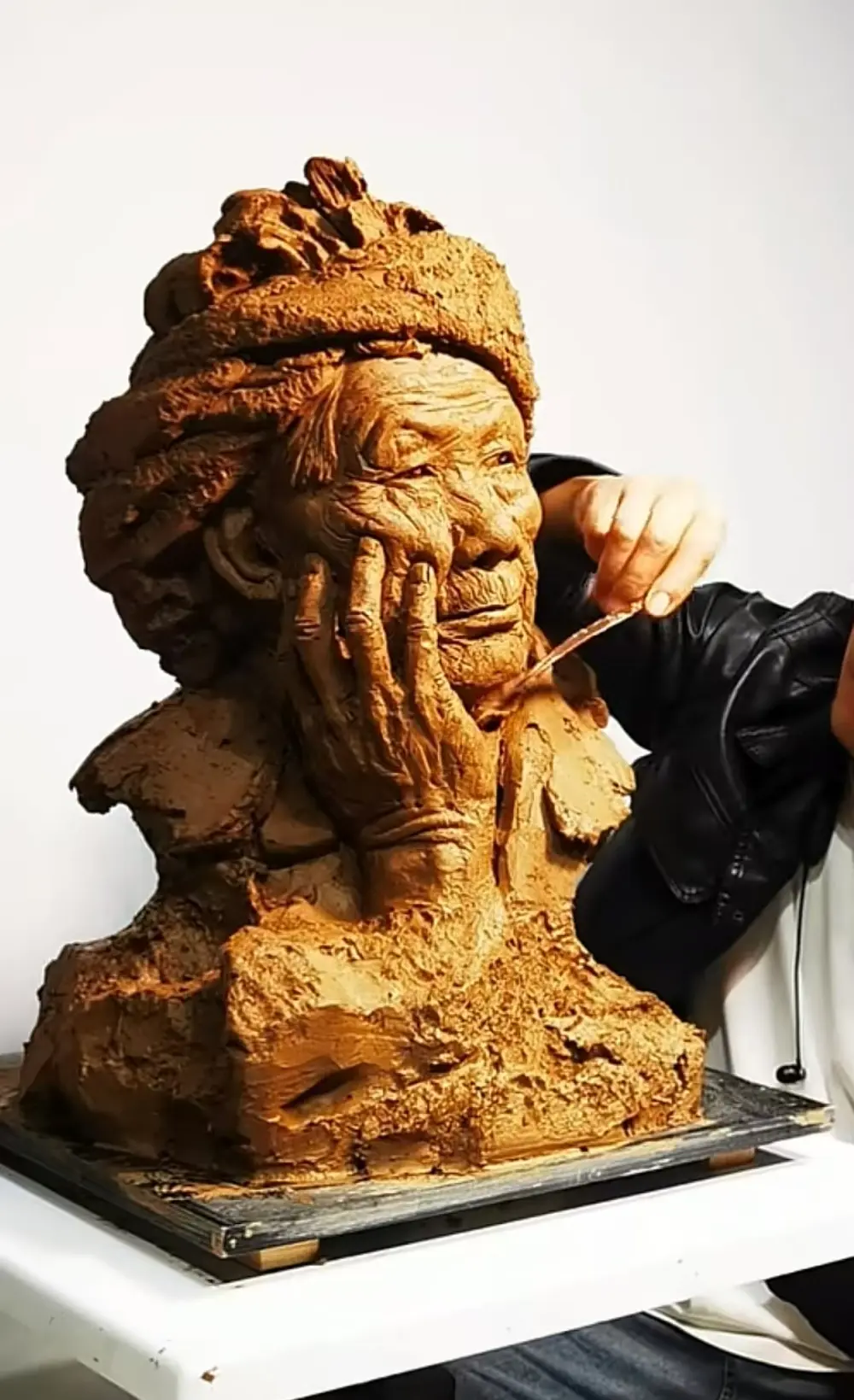 Sculptor refining the elderly lady’s clay bust in the studio, capturing the full pose, headscarf folds, and the pensive hand-to-cheek gesture