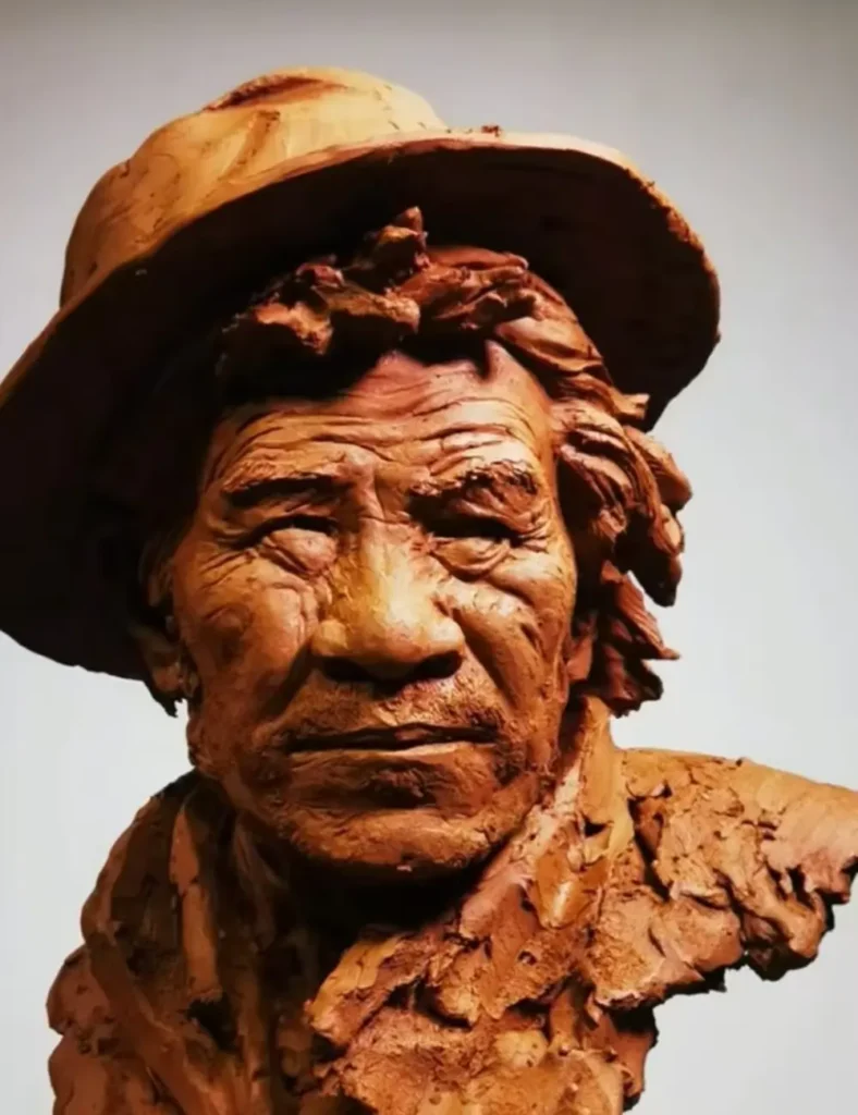 Straight-on front view of the finished clay memorial bust, capturing the elderly man’s quiet gaze, hat details, and weathered features