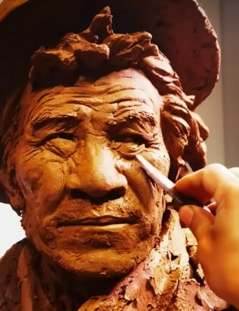 Close-up of the sculptor’s hands working on the clay bust, carefully defining the wrinkles, eyes, and hat texture for lifelike accuracy